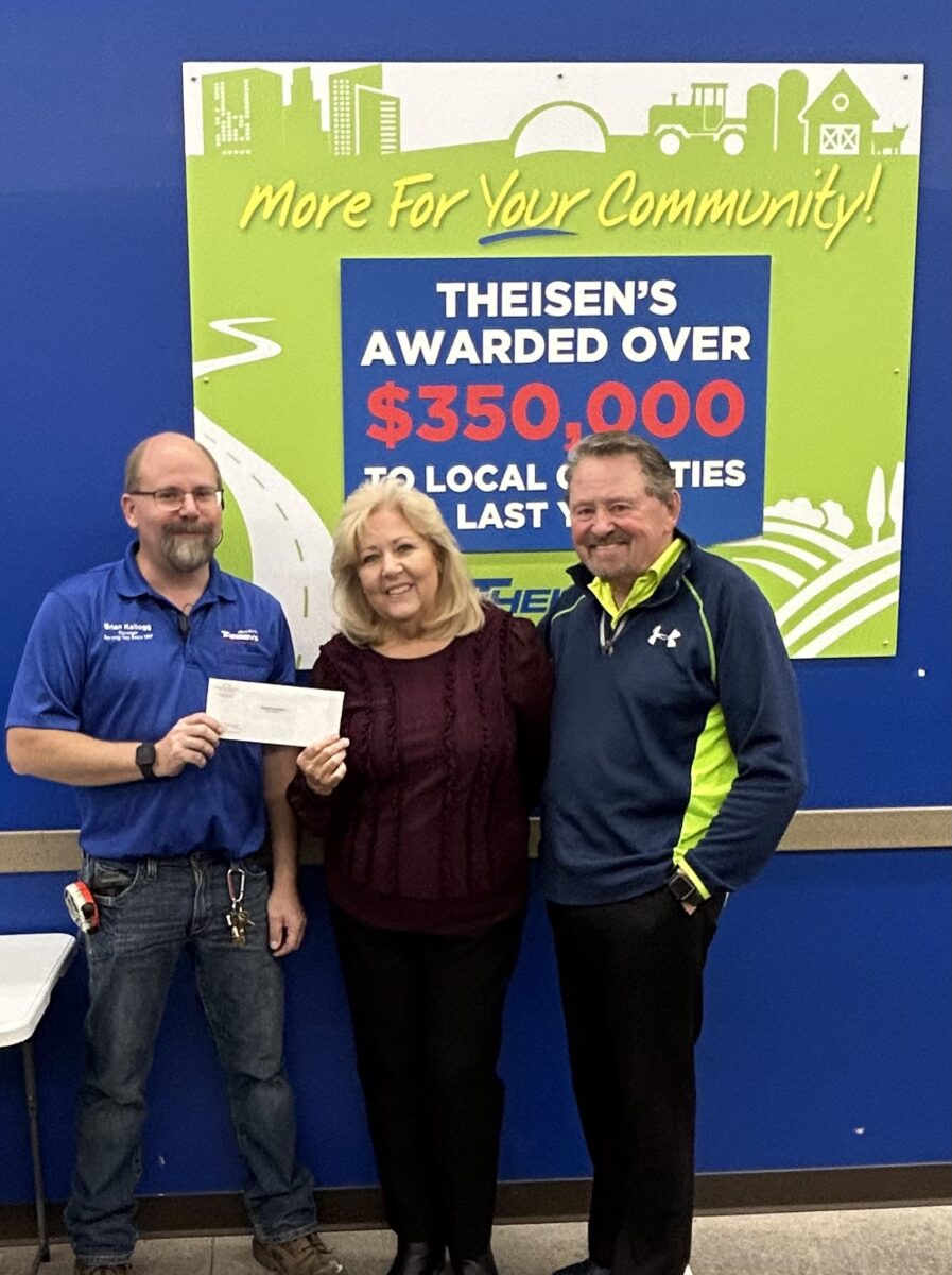 Three people smiling, with one holding a check. They stand in front of a sign promoting community donations from Theisen's.