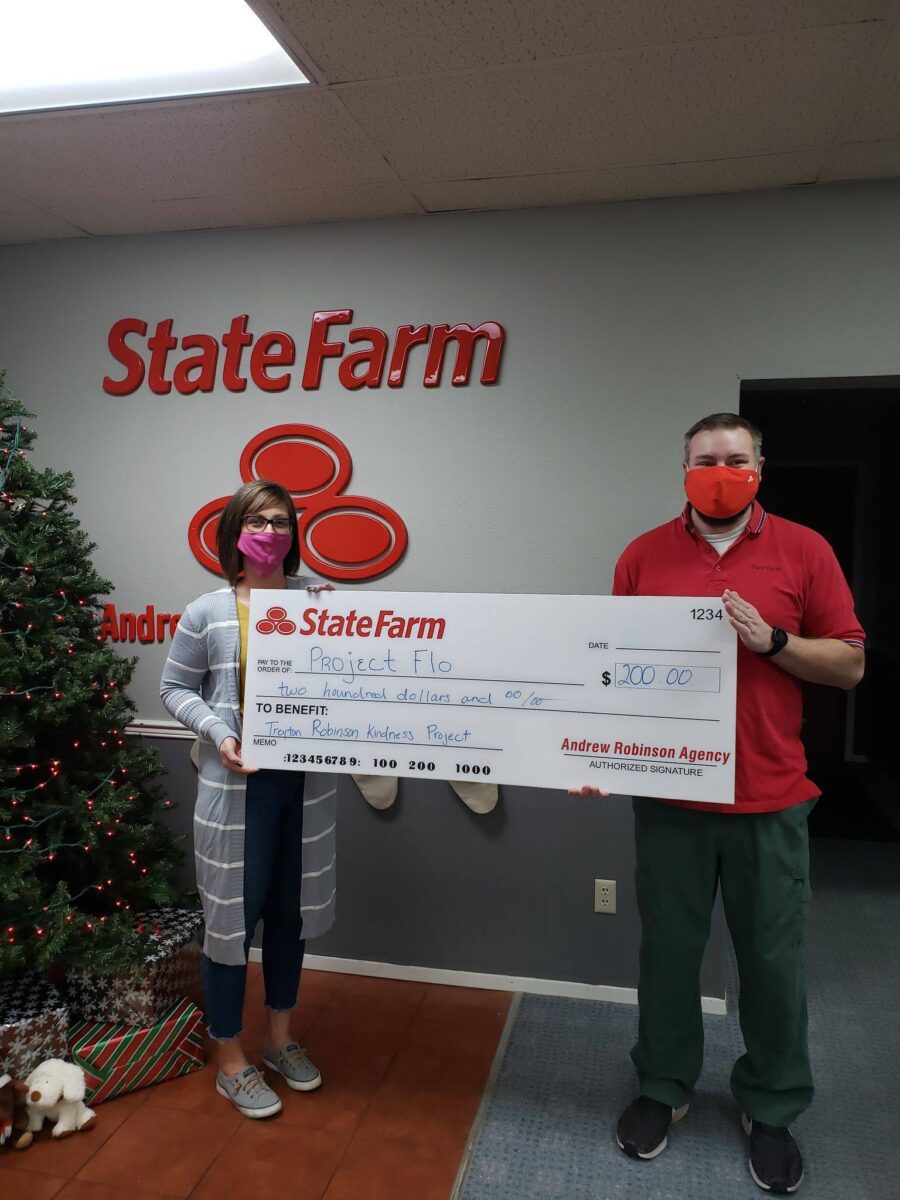 Two people holding a large check for $100 from State Farm to Project Flo, standing indoors beside a decorated Christmas tree.