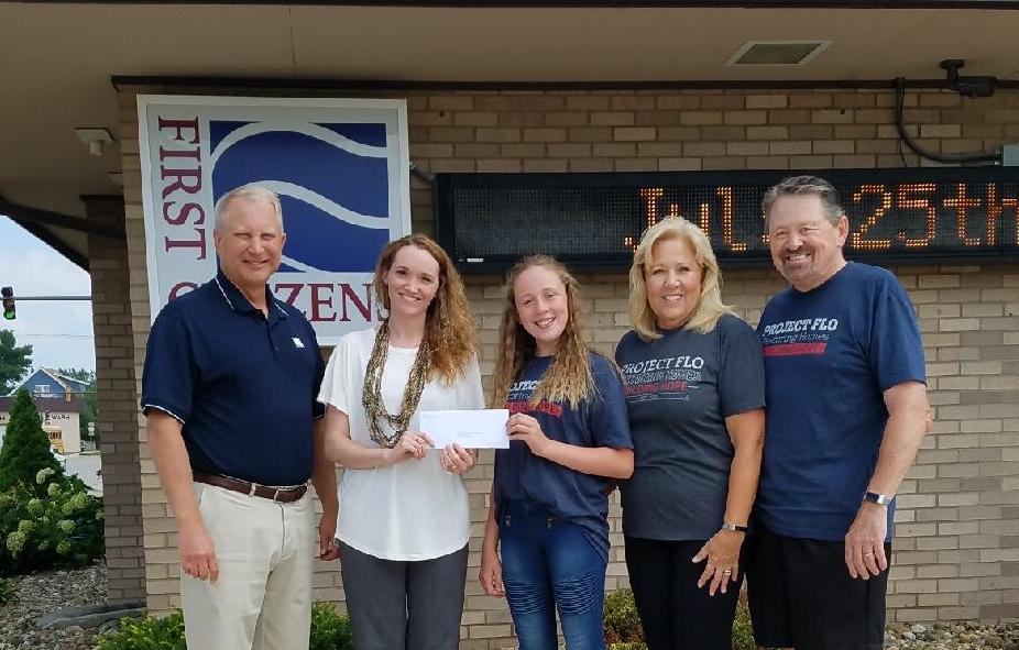 Five people standing outside a building with a sign, holding a check.
