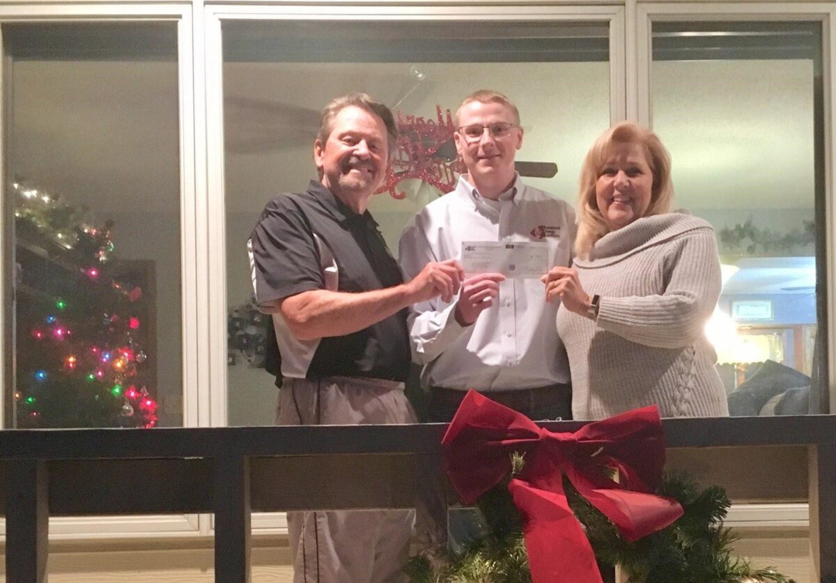 Three people stand on a porch holding a check. There's holiday decor, including a red bow and Christmas lights, in the background.