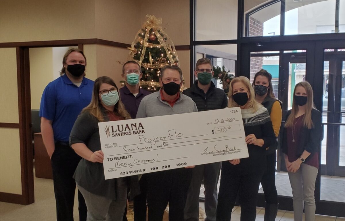 A group of people holding a large ceremonial check for Project Flo from Luana Savings Bank. A Christmas tree is in the background.
