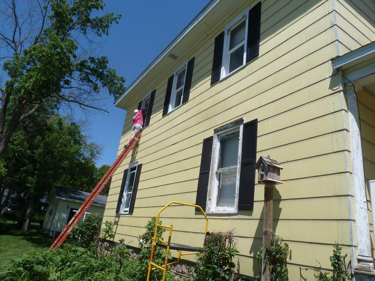 Person on a ladder painting the upper exterior wall of a yellow house with black shutters. Birdhouse on a post nearby. Bright day with clear blue sky.